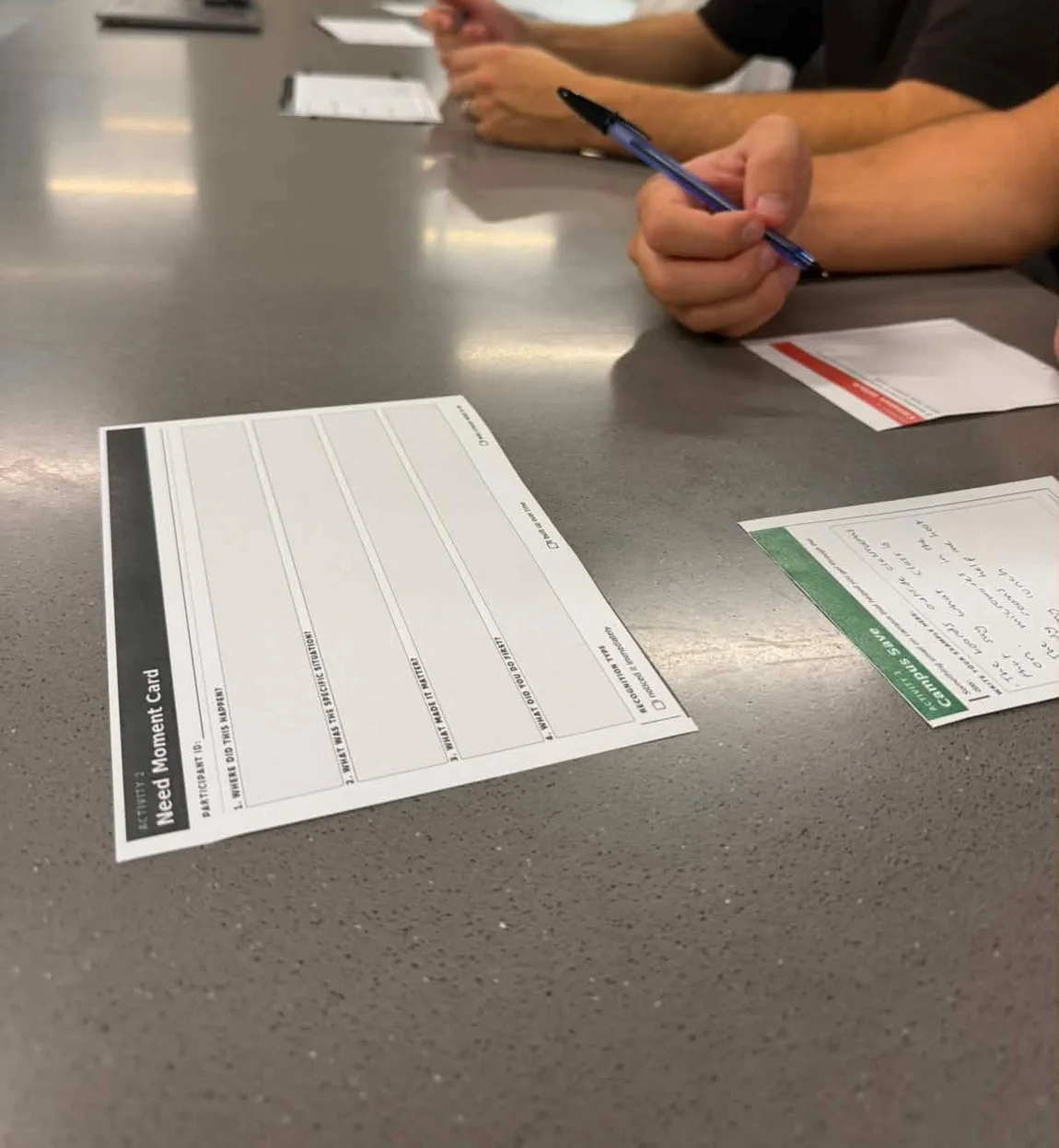 A blank Need Moment Card on the table at the start of a session, beside a Campus Save card and a participant holding a pen